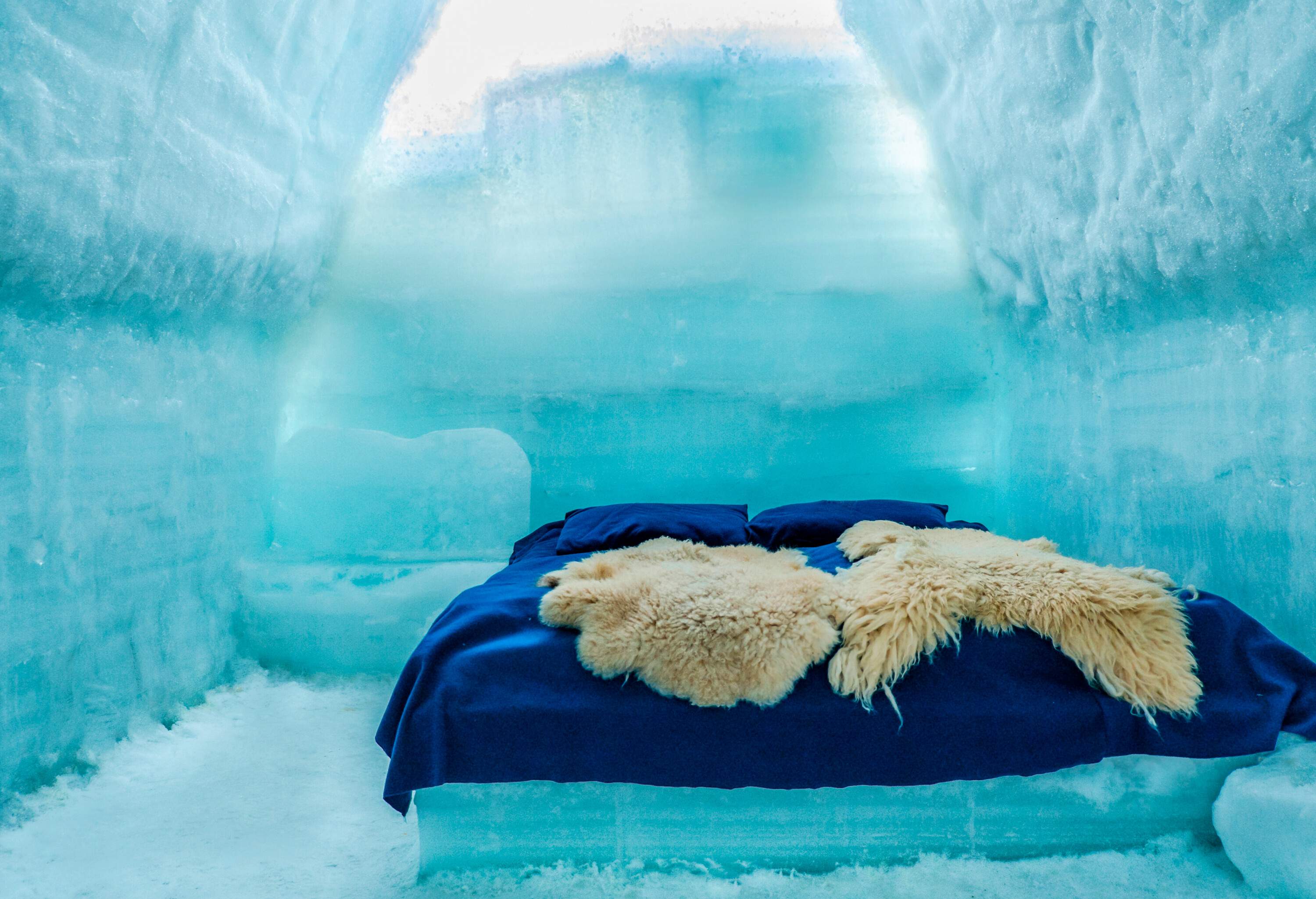 A bed inside an igloo with two beige furs, blue flat sheets and pillows.