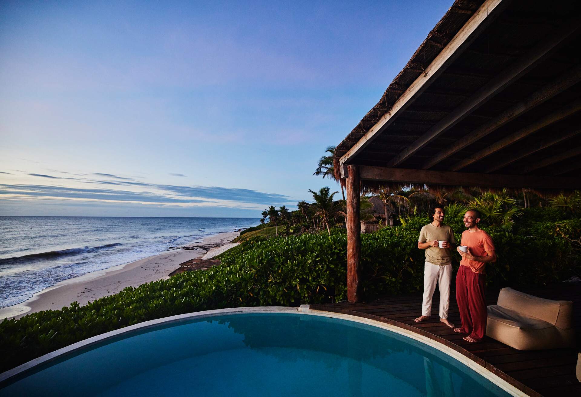 Gay couple standing poolside watching sunrise at luxury tropical villa overlooking beach and ocean, Mexico