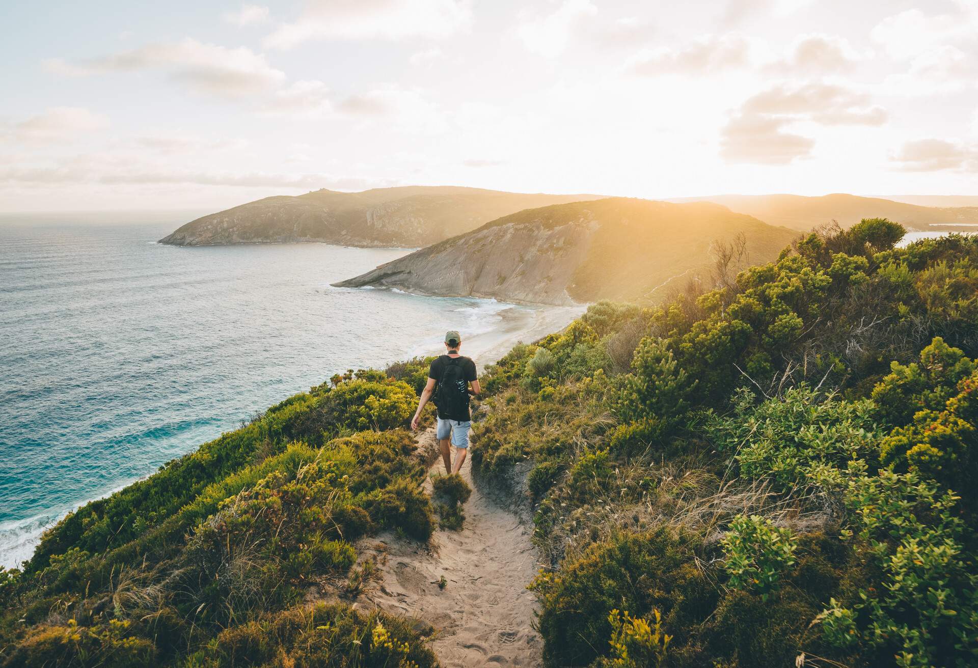 A man in a black shirt strolls down a grassy hillside path with views of the ocean and coastline.