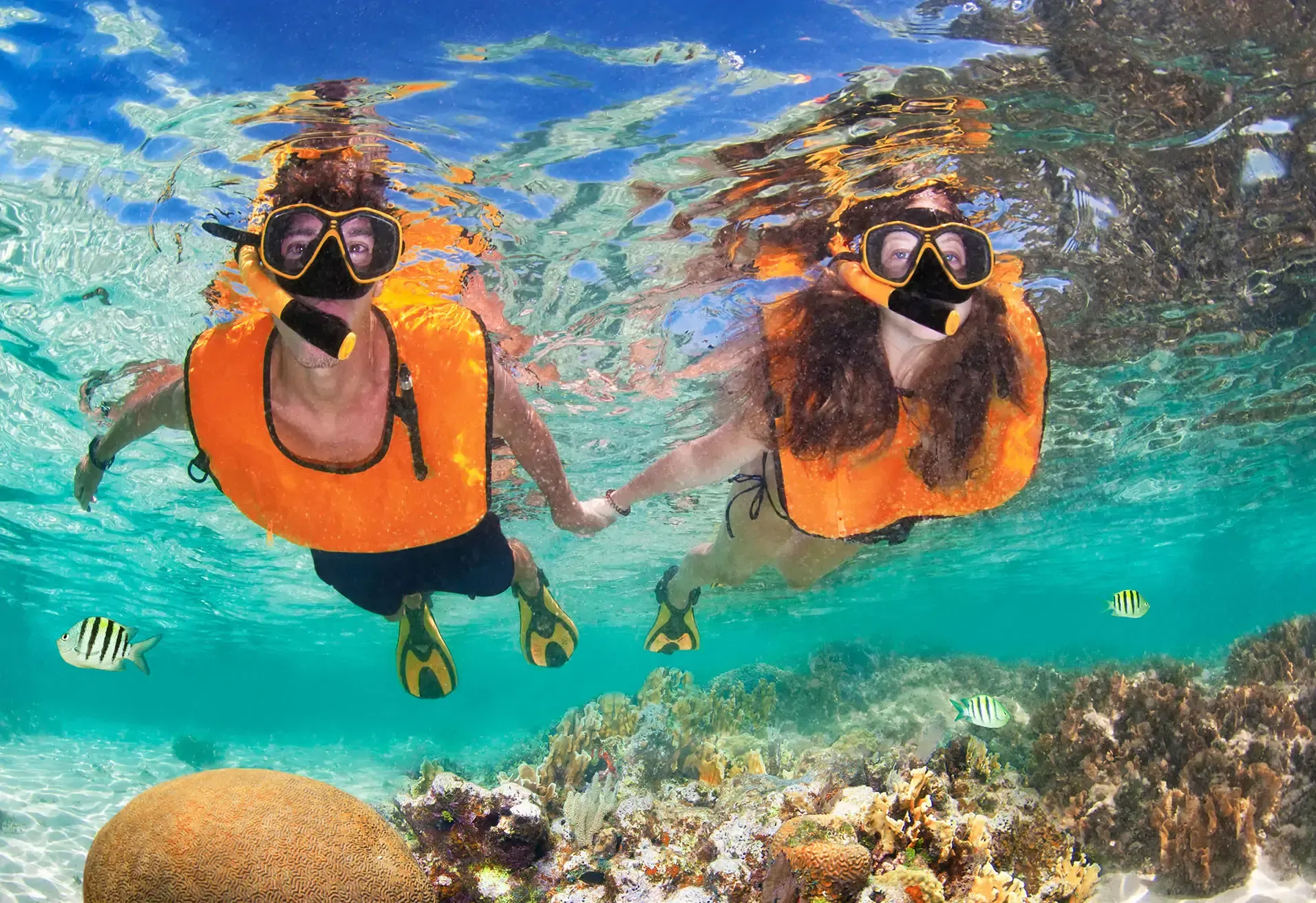 Couple snorkeling in clear tropical water, holding hands near coral reef
