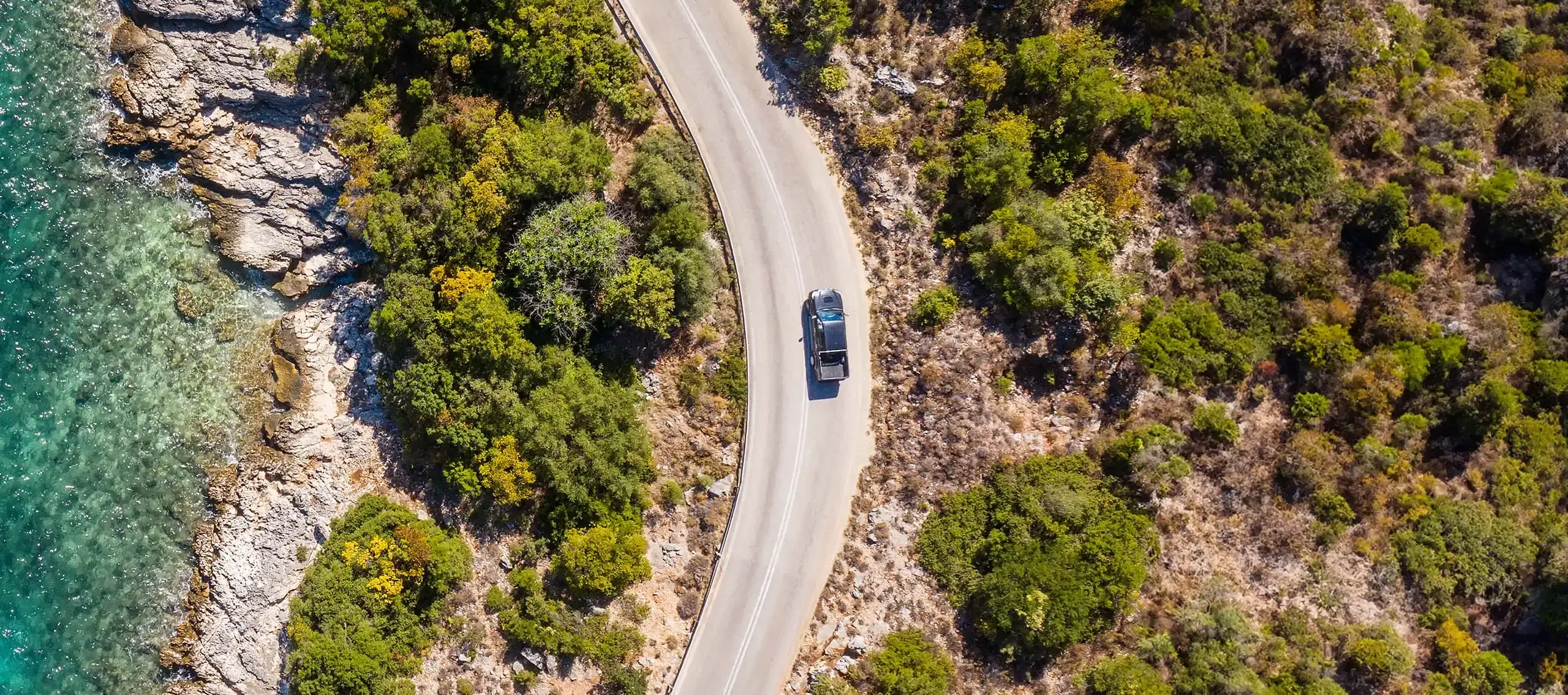 A car drives along a winding mountain road surrounded by greenery and rocky terrain.