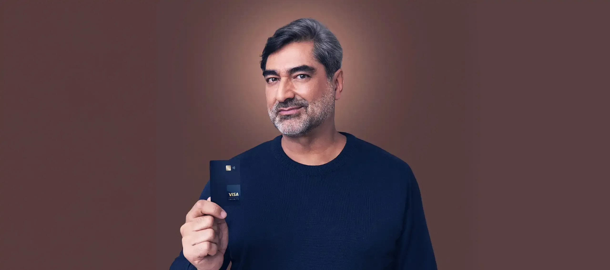 Smiling man proudly holding a Visa Infinite card against a warm brown background.