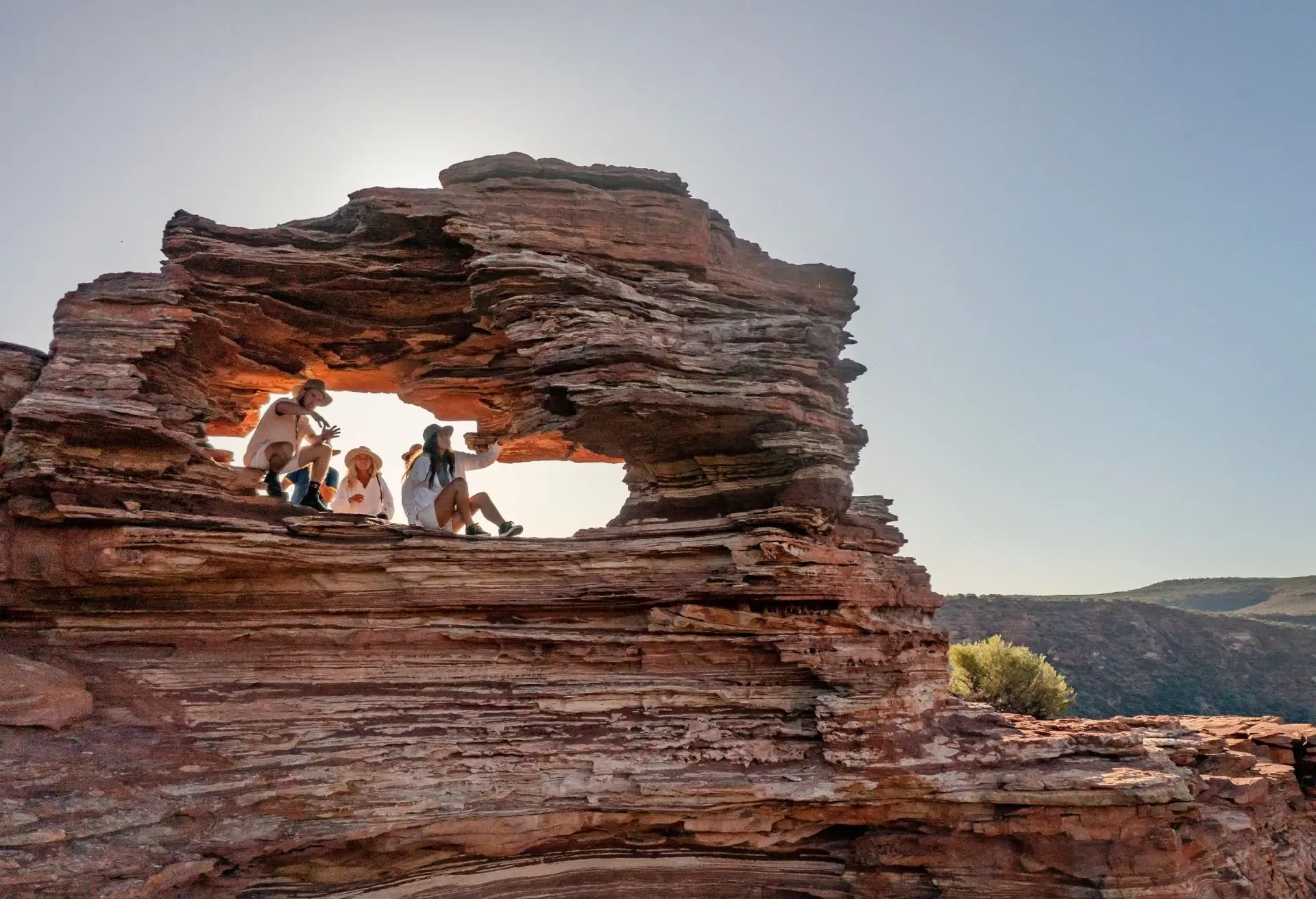 A group of people sitting inside a scenic natural arch made of layered red rock.
