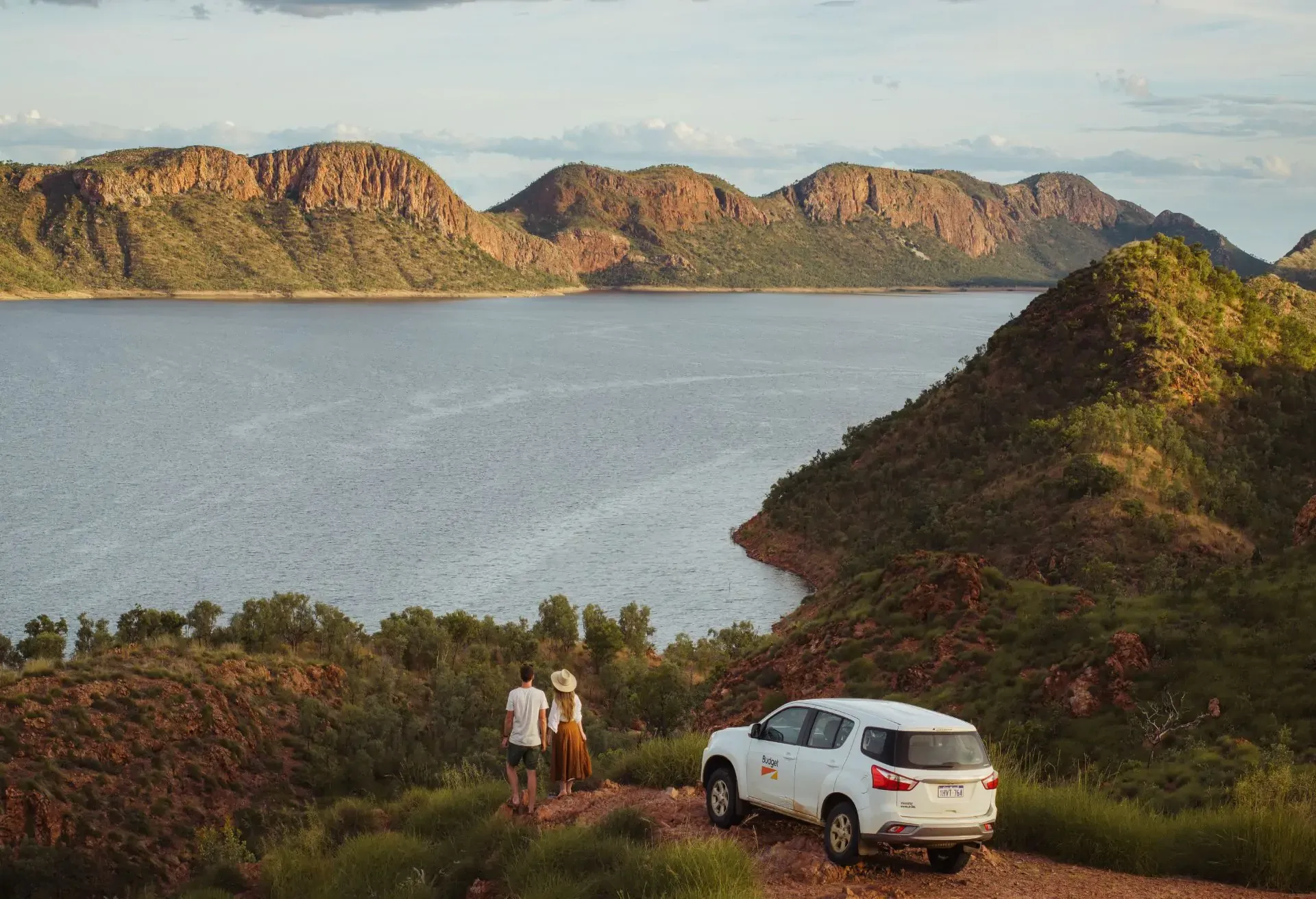 A white SUV and two people overlook a vast lake surrounded by dramatic, rocky hills at sunset.