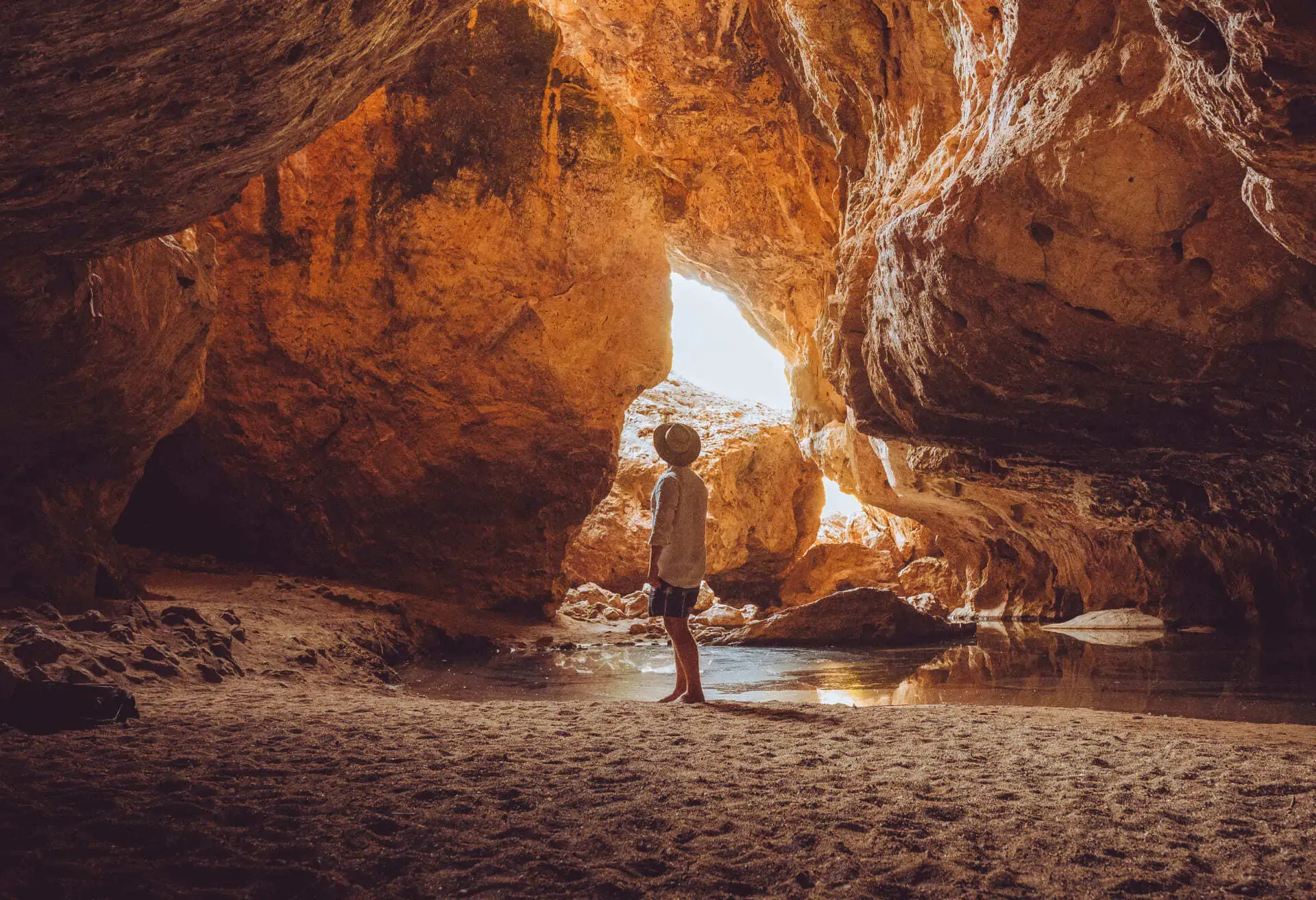 A person wearing a hat stands inside a large, sandy cave with a pool of water, lit by sunlight.