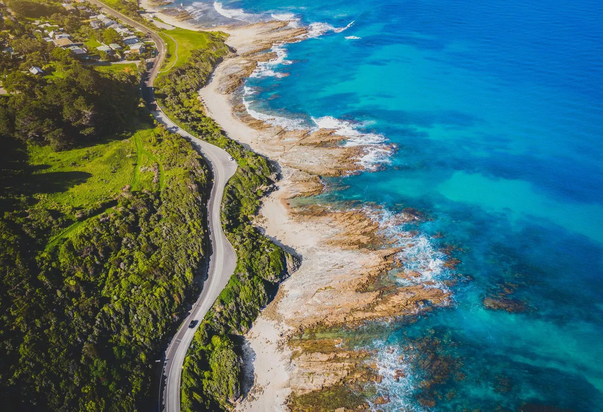 A car travels along a coastal road between lush green landscape and a rugged beach.