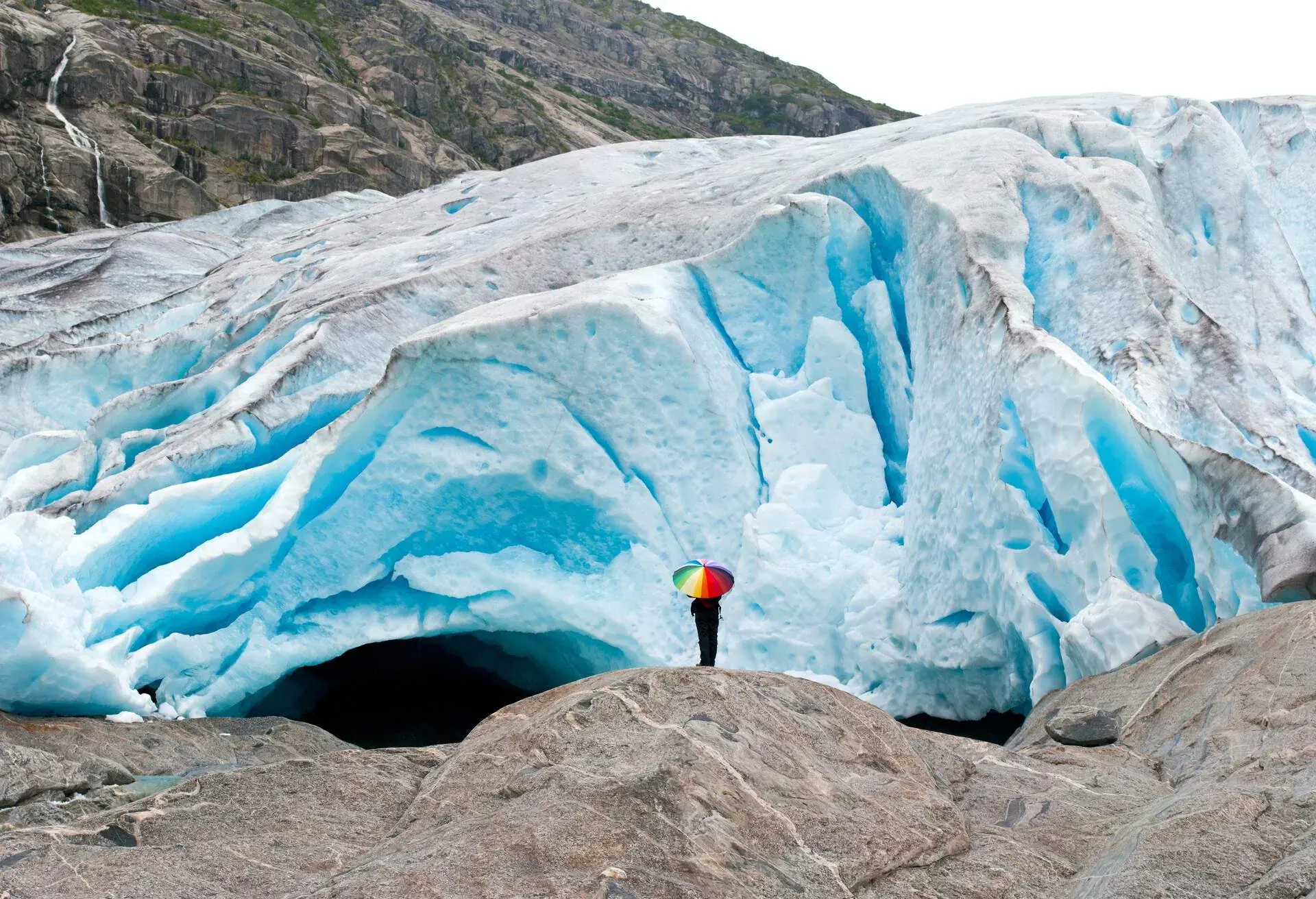 Young woman with colorful umbrella standing in front of fantastic blue glacier Nigardsbreen in Norway.