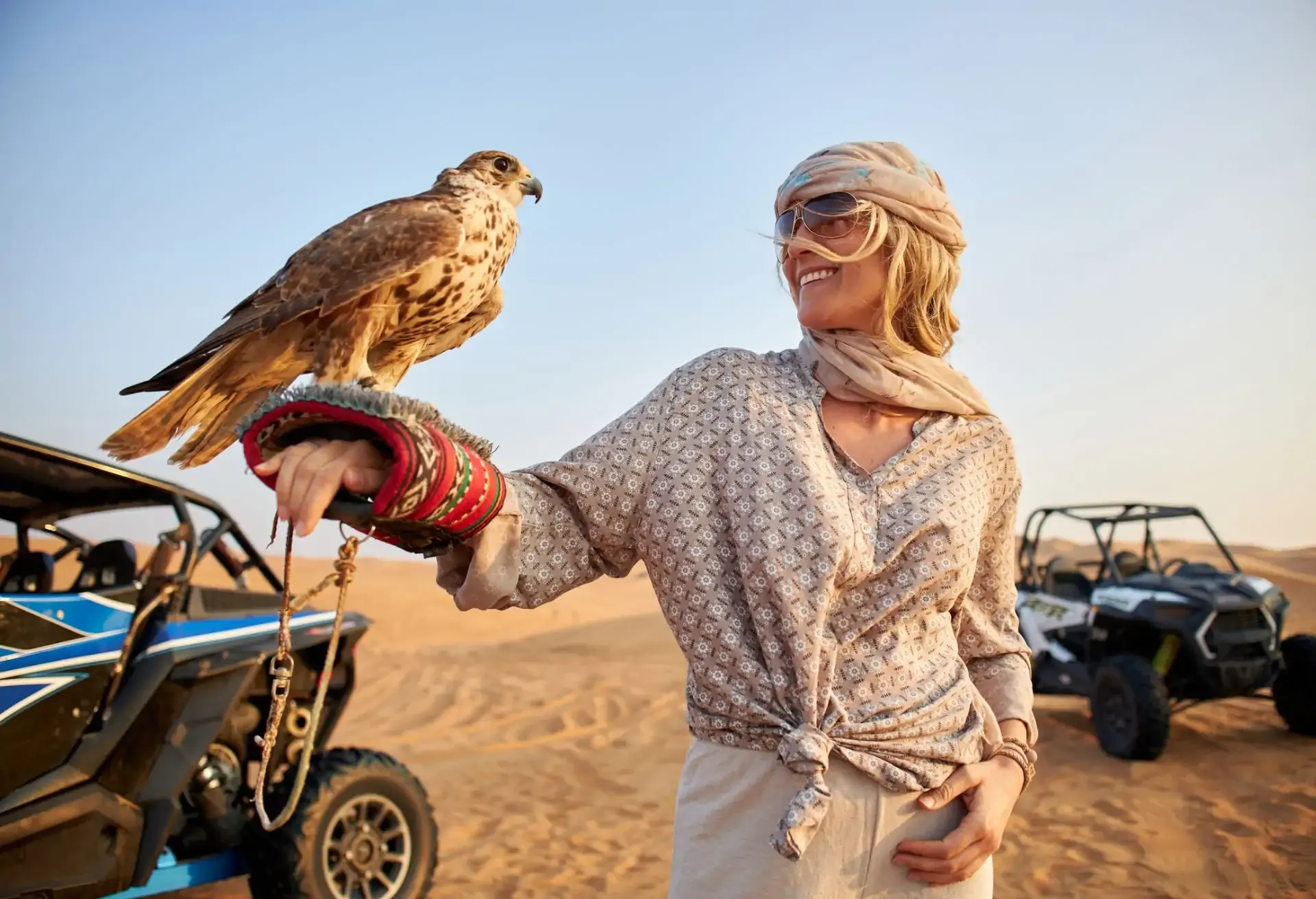 Woman smiling in desert attire holding a falcon, surrounded by desert buggies.