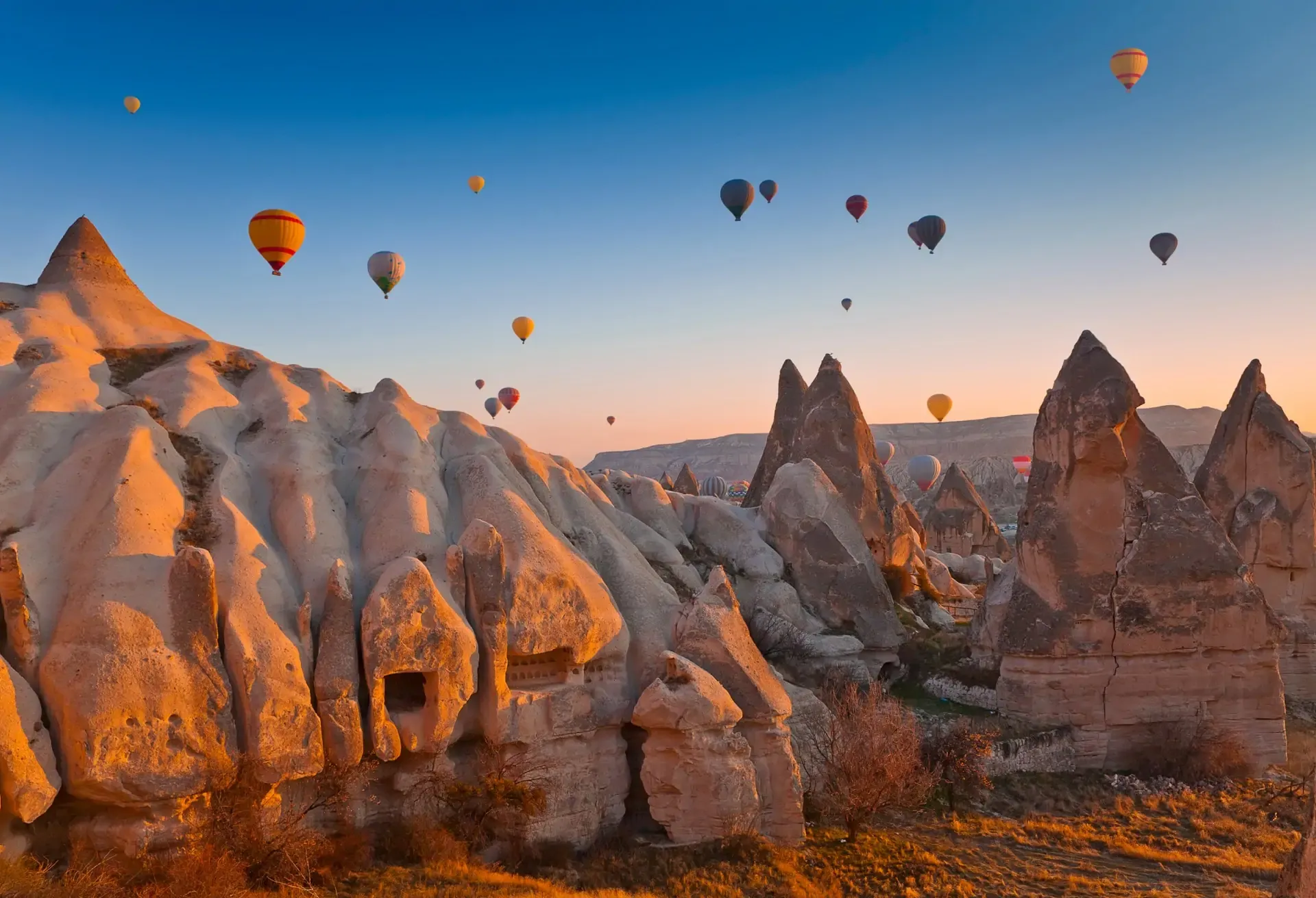 Colorful hot air balloons float over the ancient stone formations of Cappadocia during a soft sunrise.
